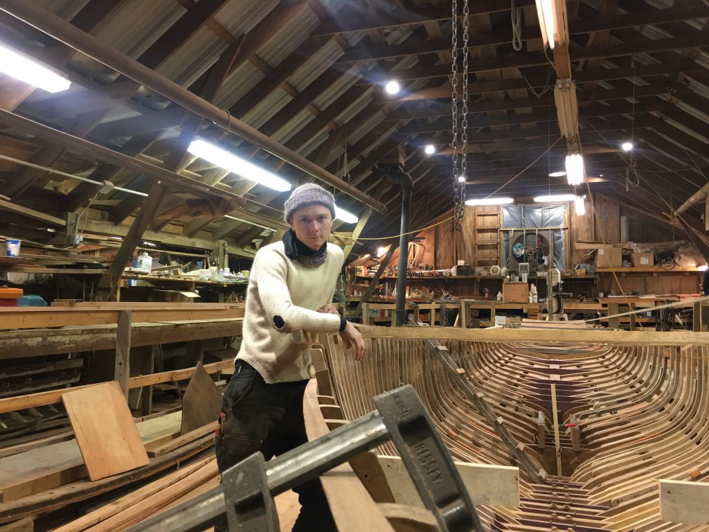 En håndværker står i et rummeligt bådværksted med træpaneler, omgivet af båddele og træbearbejdningsværktøj, og fokuserer intenst på kameraet.