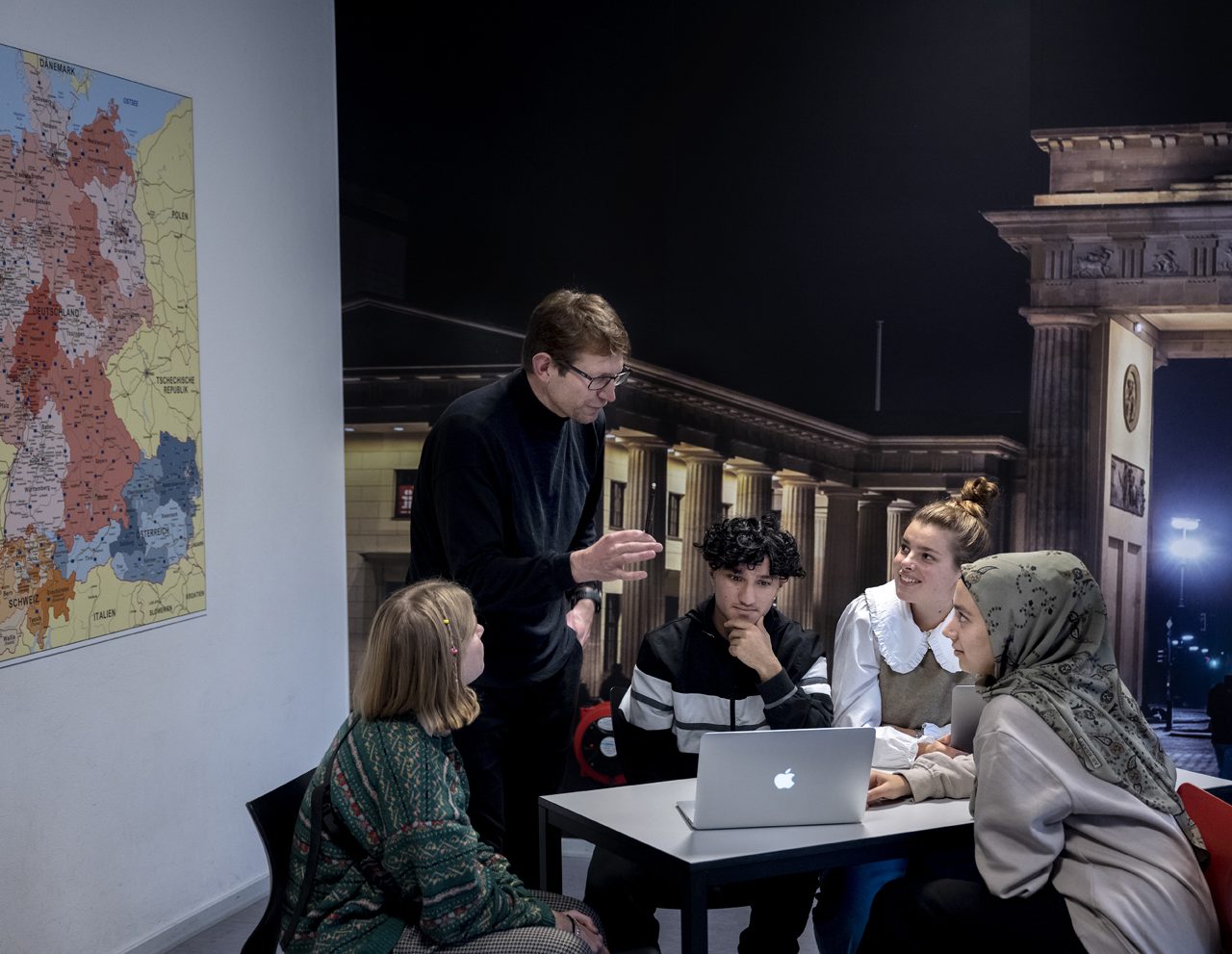 En mangfoldig gruppe på fem personer, inklusive en foredragsholder, engagerede sig i en livlig diskussion ved et bord med en bærbar computer i et rum med et kort over Tyskland på væggen og udsigt til en neoklassisk bygning udenfor.