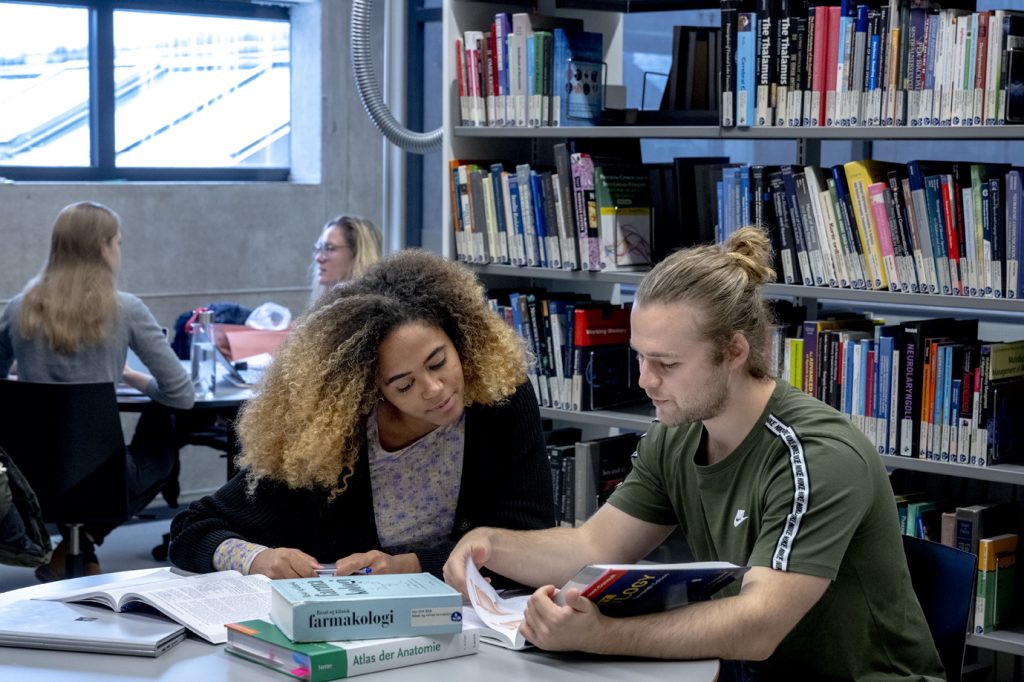 To studerende, en kvinde og en mand, studerer sammen på et bibliotek med åbne bøger og sedler på bordet. hylder fyldt med bøger og andre elever i baggrunden.