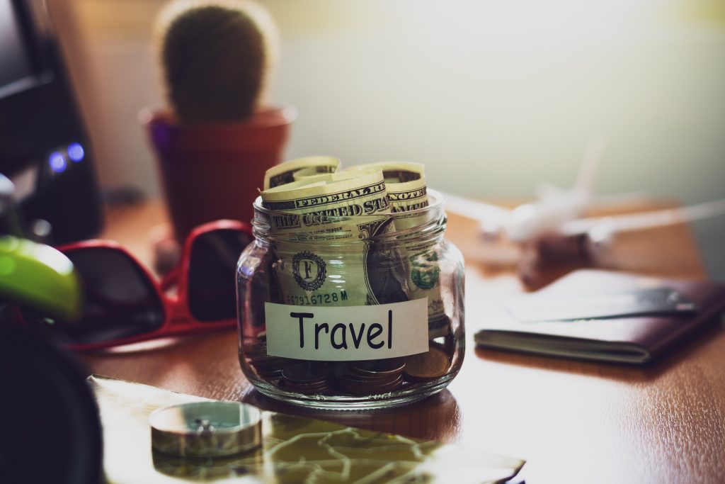 En glaskrukke fyldt med u.s. dollarsedler mærket "rejse" på et rodet skrivebord, ledsaget af en kaktus, solbriller og forskellige ting i et varmt oplyst rum.