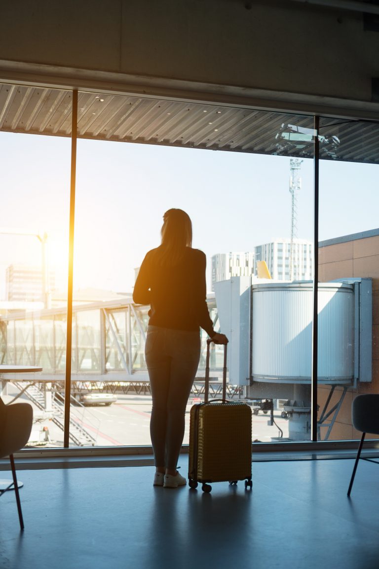 En kvinde med langt hår står i en lufthavnsterminal, oplyst af en skarp sol, holder en kuffert og kigger ud over landingsbanen.