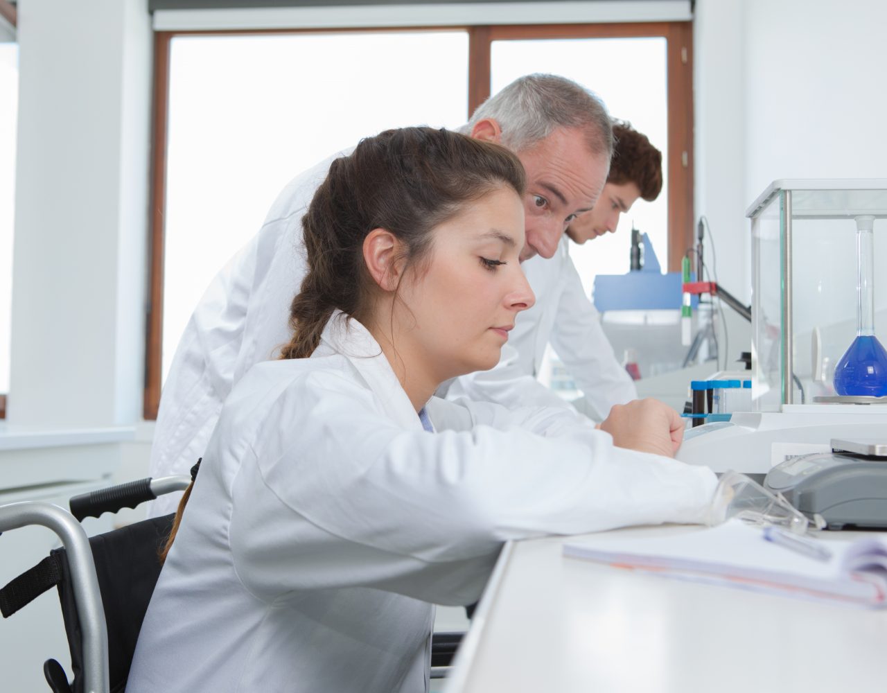En kvindelig videnskabsmand i kørestol arbejder i et laboratorium og fokuserer på sit eksperiment, som en mandlig kollega bag hende observerer opmærksomt. de bærer begge laboratoriefrakker, omgivet af laboratorieudstyr.