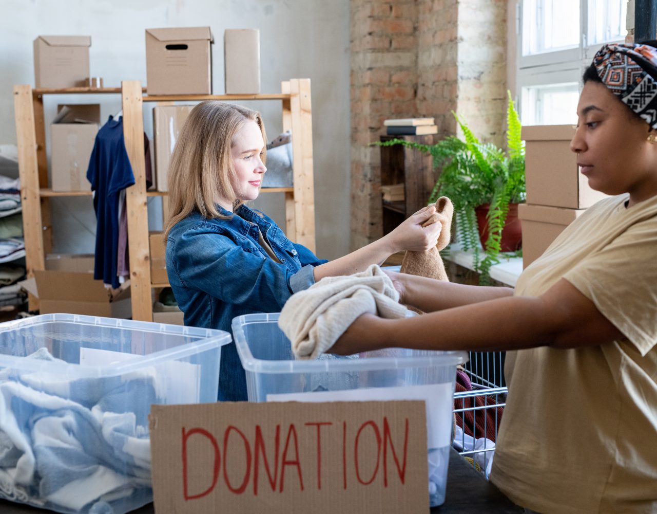 To kvinder sorterer tøj i en donationskasse i et lagermiljø og undersøger sammen et stykke tøj.