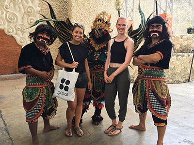 To turister, der smiler sammen med tre kunstnere i traditionelle indonesiske kostumer med kunstfærdige masker og livlige farver, sandsynligvis ved en kulturbegivenhed eller et turiststed.
