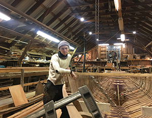 En person i et værksted står ved siden af træplanker med en båd under konstruktion i baggrunden. værkstedet er rummeligt med synlige træbjælker og værktøj.
