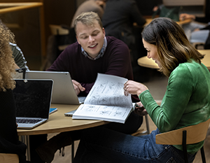 Tre kolleger diskuterer papirarbejde ved et bord med bærbare computere åbne i et godt oplyst indendørs miljø, der udviser engagement og teamwork.
