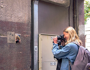 En kvinde med et kamera tager et billede af et lille indrammet billede af et ansigt, monteret på en byvæg ved siden af en metaldør.