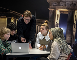 En forskelligartet gruppe på fem unge voksne, inklusive en kvinde i hijab, er samlet omkring en bærbar computer, engageret i en diskussion ledet af en stående mand, i bymiljøer om natten.
