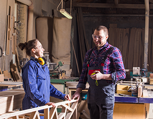 To tømrere, en mand og en kvinde, diskuterer en træramme i et værksted fyldt med værktøj og trærester. manden holder et målebånd, og begge smiler.