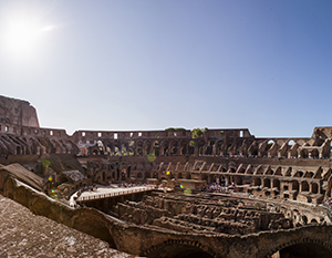 Interiør af Colosseum i Rom, der viser dets enorme elliptiske struktur med overlevende etagede siddepladser og buer under en klar himmel. sollys oplyser det gamle arenagulv og stenvægge.