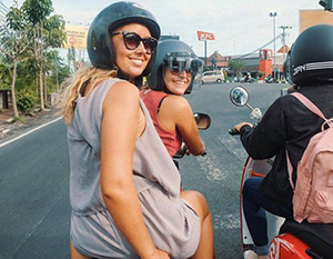 To kvinder med hjelme smiler, mens de sidder på en scooter på en travl bygade.
