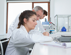 Tre videnskabsmænd, to kvinder og en mand, der arbejder i et laboratorium. de er fokuseret på at skrive og gennemgå noter. laboratorieudstyr er synligt i baggrunden.