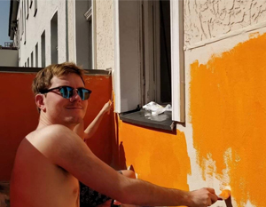En mand med solbriller maler en udendørs væg orange og smiler til kameraet på en solskinsdag.