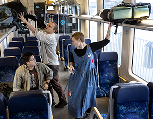 Tre personer i en togvogn: en mand, der stuver bagage over hovedet, en anden, der sidder og kigger op, og en stående kvinde, der justerer en strop på hendes overalls.