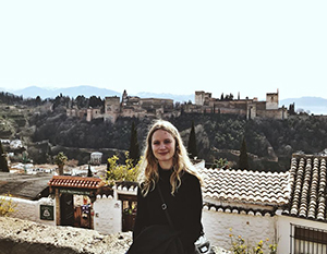En kvinde smilende i forgrunden med alhambra-paladset og fæstningskomplekset i Granada, Spanien, synligt i baggrunden, sat mod en klar himmel.