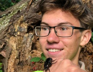 En teenagedreng med briller, der smiler, mens han holder en stor sort bille på hånden, med en naturlig baggrund med grønt og en træstamme.