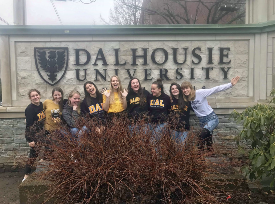 Otte unge kvinder smiler og poserer foran et "dalhousie university"-skilt, nogle iført universitetsmærket tøj.