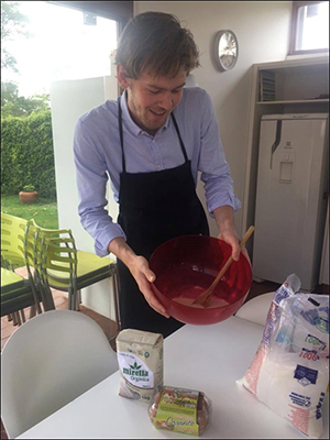En ung mand iført et mørkt forklæde blander entusiastisk ingredienser i en stor rød skål i et køkken med udsigt til haven. ingredienser som mel, smør og yoghurt er synlige på bordet.