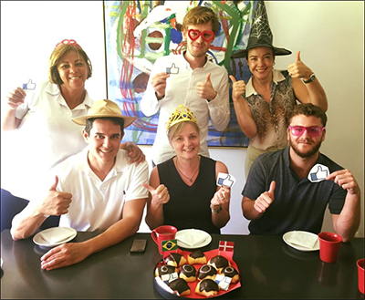 Syv personer poserer ved en festlig sammenkomst, giver tommelfingeren op og holder papir-lignende ikoner, iført forskellige hatte, med en tallerken desserter på bordet.