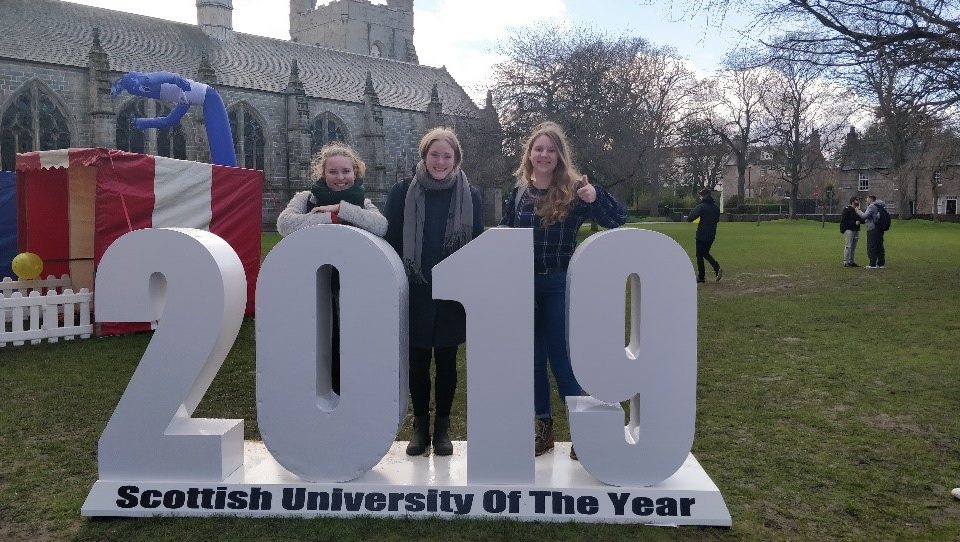 Tre kvinder, der smiler og står bag store "2019"-tal ved en begivenhed, der fejrer "årets skotske universitet" på en solskinsdag med græs, træer og mennesker i baggrunden.