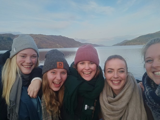 Fem kvinder smilende i en selfie ved en sø med bakker i baggrunden, iført vinterhuer og tørklæder.