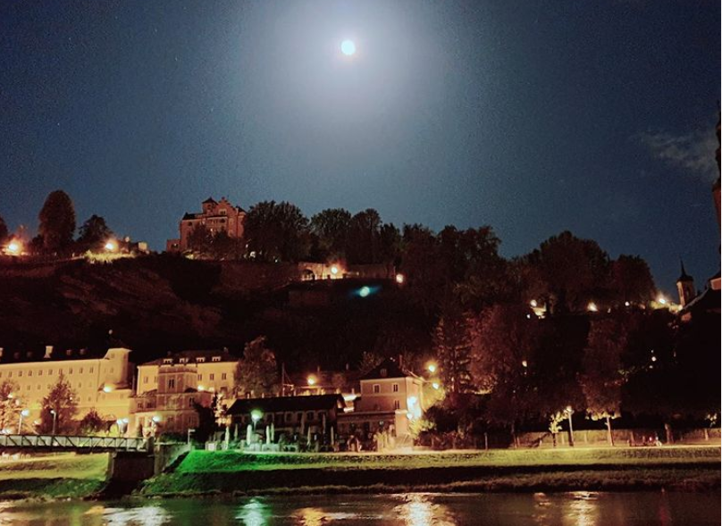 Natteudsigt over en oplyst by ved floden med et slot på en bakke under fuldmåne. skarpe lys reflekterer fra vandet og tilføjer en varm, rolig glød til scenen.