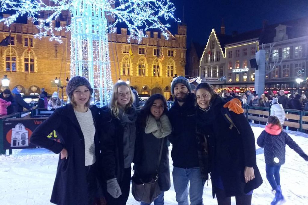 Fem venner poserer glade foran et oplyst juletræ og festlige lys på et travlt udendørs vintermarked på en historisk plads.