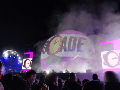 Publikum ser en levende koncert om natten med en stor oplyst scene, der viser teksten "arcade" og stiliseret urgrafik, omgivet af røg og farvet lys.