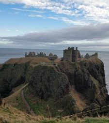 En naturskøn udsigt over dunnottar-slottet, der ligger på toppen af en forrevne klippe med udsigt over havet, med en overskyet himmel over og en græsklædt sti, der fører til ruinerne.